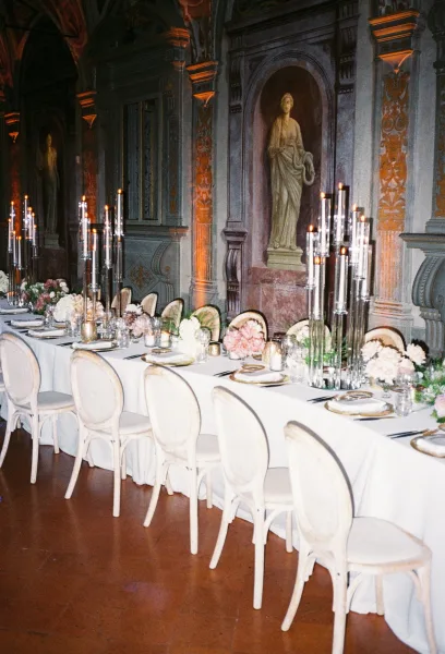 Reception tablescape with long banquet table decor, set with taper candles, beneath fresco walls and classical statues amid arches.