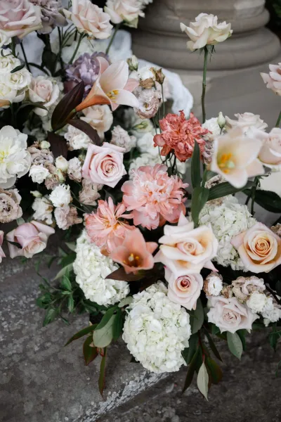 Wedding floral arrangement of blush and peach wedding flowers with roses, styled along stone steps beside a column draped in white fabric.
