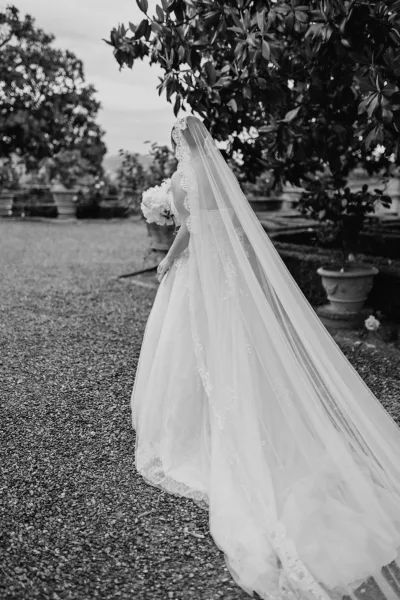 Bridal portrait of a bride with cathedral veil sweeping along a gravel path, framed by garden trees and potted plants