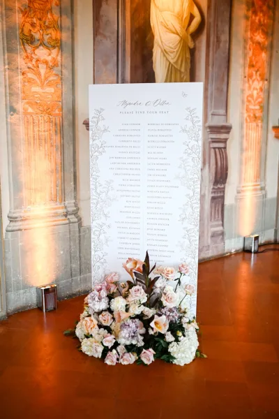 Wedding seating chart serving as a find your seat sign, edged with roses, set before an ornate mural wall and classical statue under uplighting.