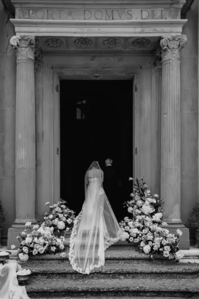 Ceremony entrance as a bride entering church ascends stone steps toward a church doorway, bridal veil trailing, framed by stone columns.