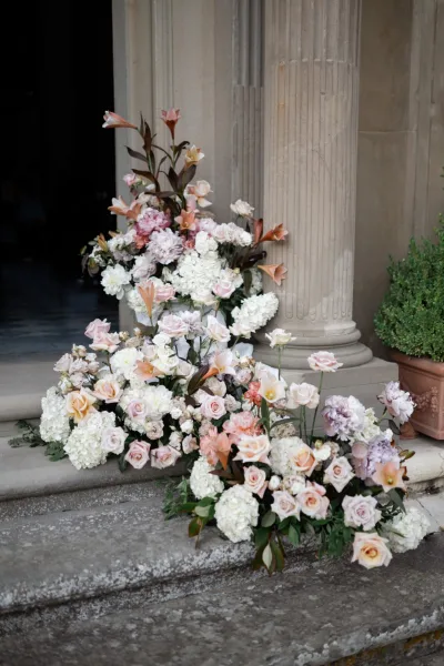 Wedding entrance flowers arranged in lush clusters with roses, lining stone steps and a column at a stone doorway with potted boxwood.