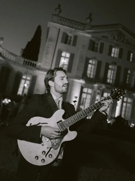 Wedding guitarist playing electric guitar, delivering live wedding music on a garden lawn before a lit mansion under the night sky.