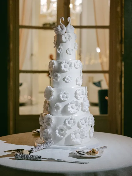 Wedding cake, a sugar flower wedding cake with a graceful swan topper, stands tall before window panes and glowing chandeliers.