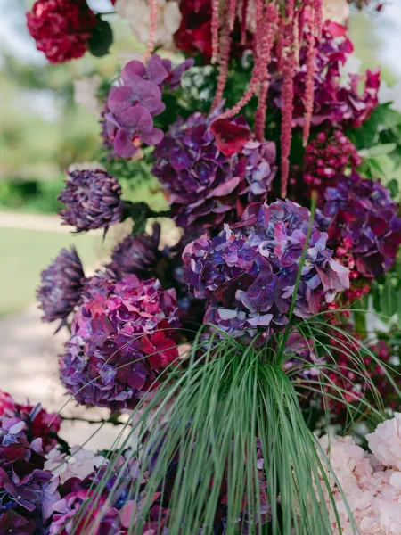 Floral arrangement of purple wedding flowers with trailing amaranthus, set against a lush garden lawn beside a winding stone pathway.