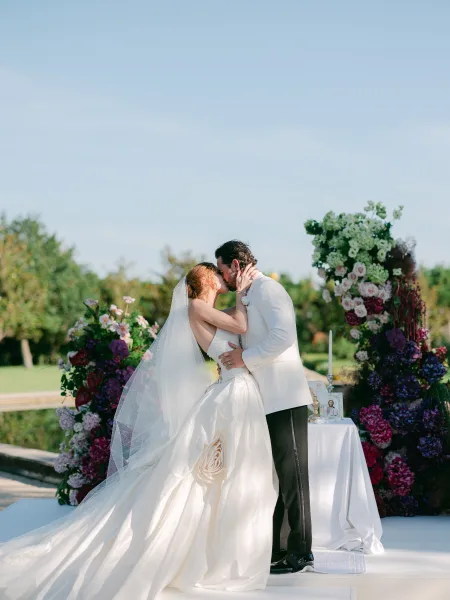 Wedding first kiss in a garden ceremony moment, framed by floral pillars beside a reflecting pool with garden trees under a blue sky.