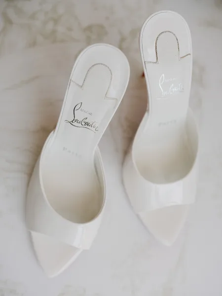 Bridal shoes—white bridal mules with pointed toes—styled neatly on a subtly textured white surface.