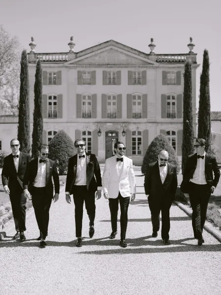 Groom and groomsmen walk a gravel path, the groom in white tux leading, bow ties sharp, framed by a manor house and tall cypress.