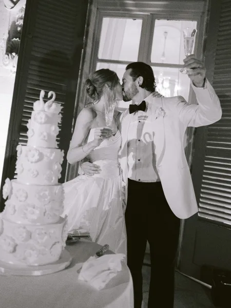 Wedding champagne toast as a couple kiss at the reception beside the wedding cake, framed by soft drapery and chandelier light