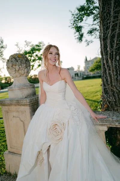 Bridal portrait in a strapless wedding dress with floral appliqué, posed on a garden lawn near a stone sculpture and historic estate.