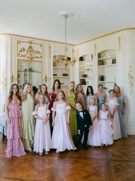 Wedding party portrait with bridesmaids and flower girls, plus a ring bearer in a classic tuxedo, posed before ornate paneling and a chandelier.