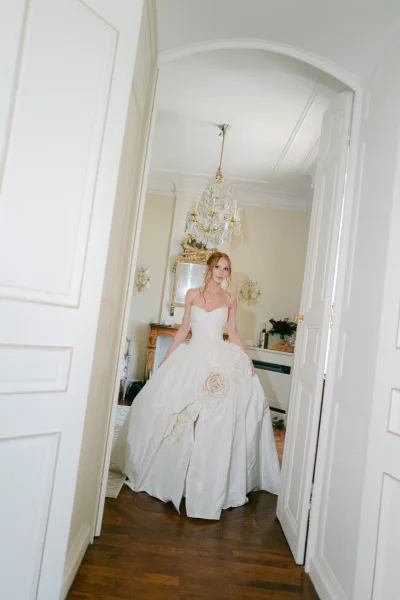 Bridal portrait of a bride in ballgown, beneath a crystal chandelier, standing in an arched doorway framed by cream walls.