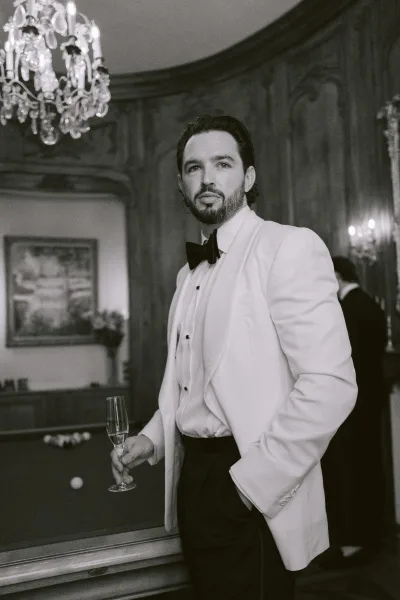 Groom portrait of a groom in white tuxedo, holding a champagne flute beside a billiards table under a chandelier.