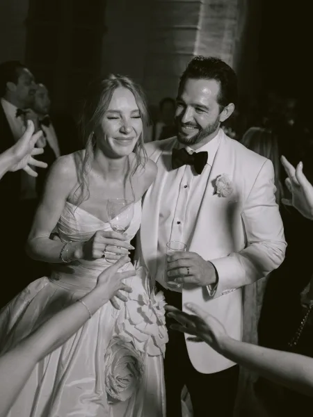 Wedding reception moment with a bride and groom laughing, holding cocktail glasses by a stone wall and shuttered window as friends cheer.