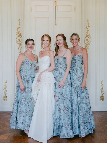 Bride and bridesmaids in matching bridesmaid dresses, the bride in a gown with floral appliqué, before paneled doors with gold moulding.
