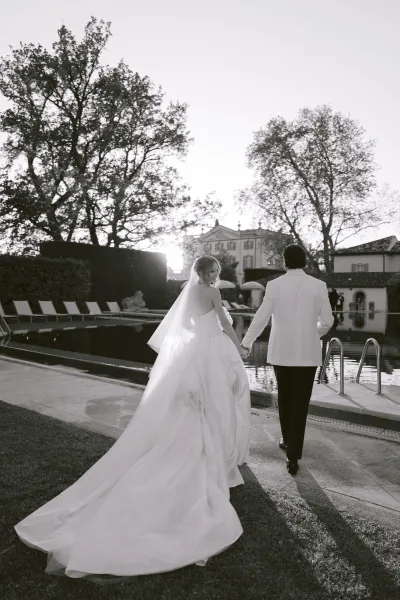 Couple portrait of the bride and groom walking hand in hand, her veil trailing, along a stone path by a pool with villa architecture and trees.