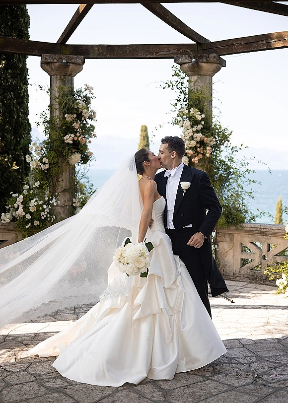 Wedding kiss portrait of bride and groom kissing under a floral arch, her long veil trailing, on a stone terrace with ocean view