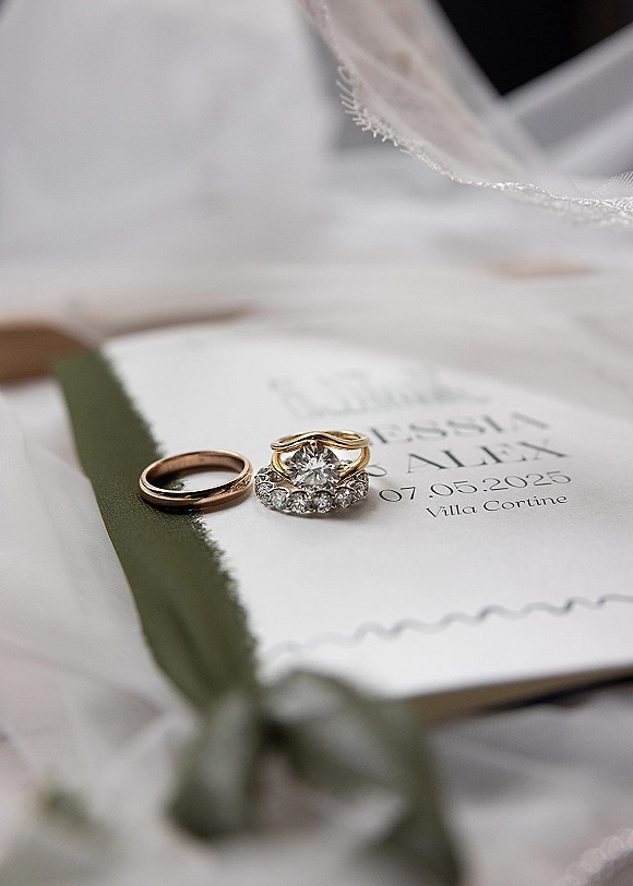 Wedding rings and engagement ring close up, diamond ring and band resting on deckle-edge invitation with sage green ribbon on white backdrop