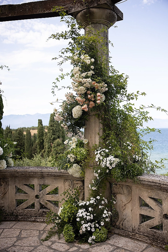 Wedding floral installation with ceremony pillar flowers, climbing greenery, roses and hydrangeas on a stone terrace overlooking lake and mountains
