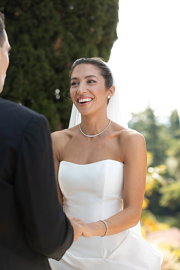 Ceremony moment as bride smiling at groom, holding his hands in a strapless wedding dress with veil, amid sunlit trees and sky