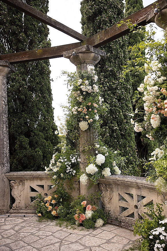 Wedding floral arch draped with ceremony altar flowers, roses and hydrangeas on stone columns under a pergola with string lights and evergreens