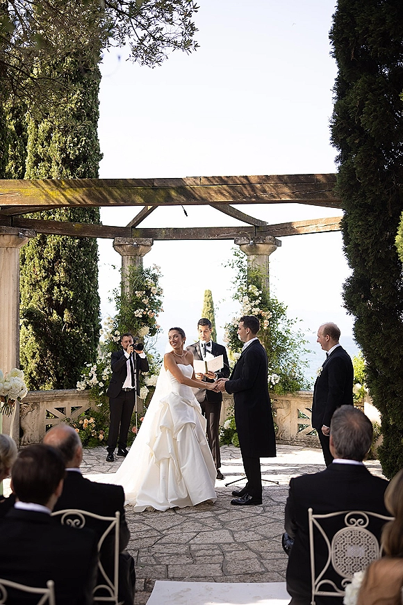 Wedding vows at an outdoor wedding ceremony as the bride and groom hold hands under a floral arch on a garden terrace with stone columns