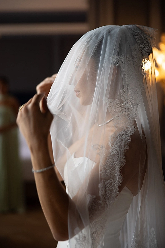 Bridal portrait of a bride with veil over face in warm window light, wearing a strapless wedding dress and lace veil in a blurred room