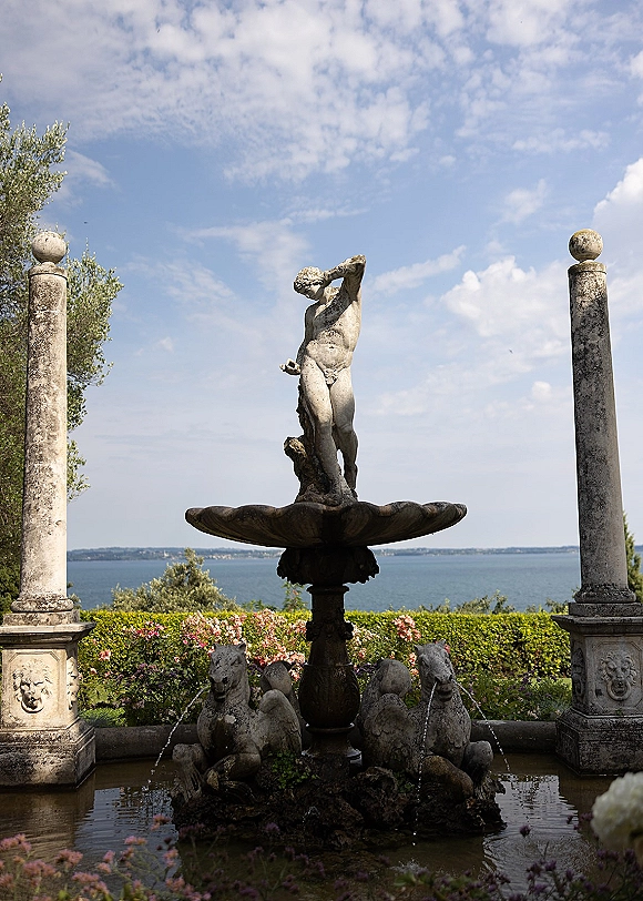 Garden fountain with stone fountain statue and water jets, framed by carved columns, flower beds, and a lake view under blue sky