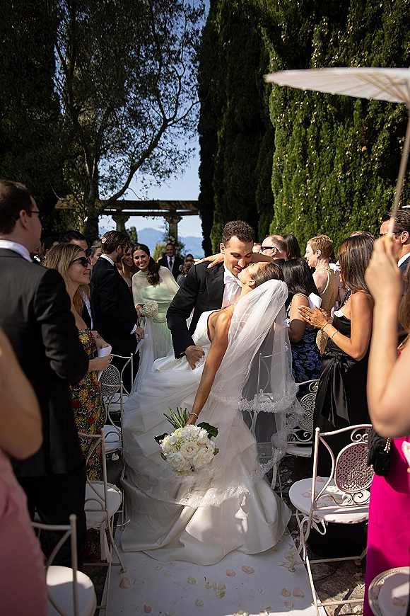 Wedding recessional as bride and groom share an aisle kiss on a white runner with rose petals, guests cheering in a hedge-lined garden