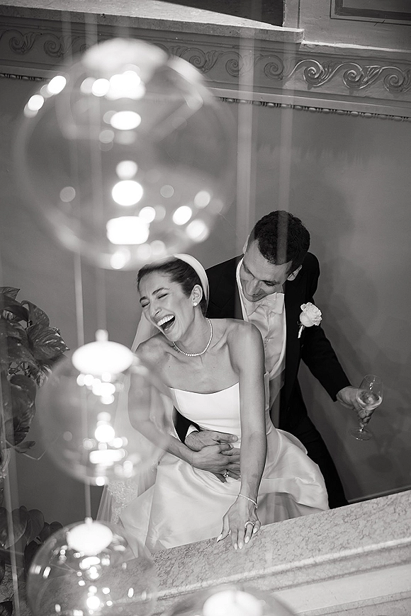 Couple portrait of a laughing bride and groom on an indoor staircase, groom hugging her as chandelier lights glow behind them