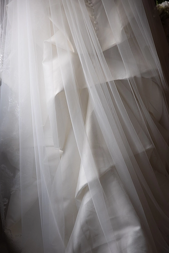 Wedding dress detail with bridal veil draped over a satin gown, highlighting fabric-covered buttons in soft window light and shadows