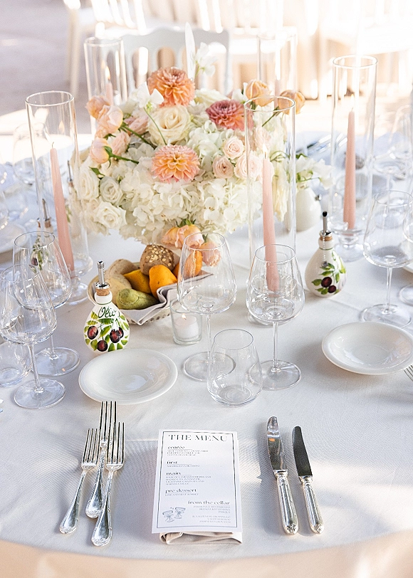 Reception tablescape with a wedding table centerpiece, blush taper candles, and peach-white florals on white linen in soft natural light