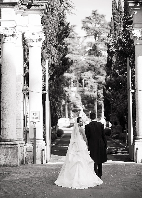 Couple portrait of newlyweds holding hands walking away, bride glancing back in cathedral veil by stone columns and garden gate path