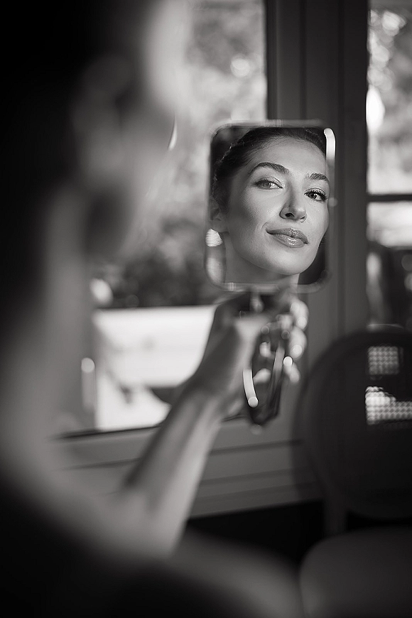 Bridal makeup close-up with soft glam lip color as the bride holds a handheld mirror, earring visible by a window in an indoor room