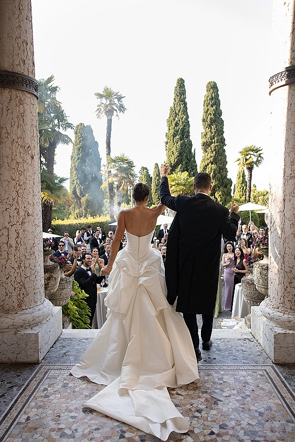 Wedding recessional as bride and groom exit with raised hands, her strapless gown’s long train trailing on mosaic floor past cheering guests on a garden terrace with palms