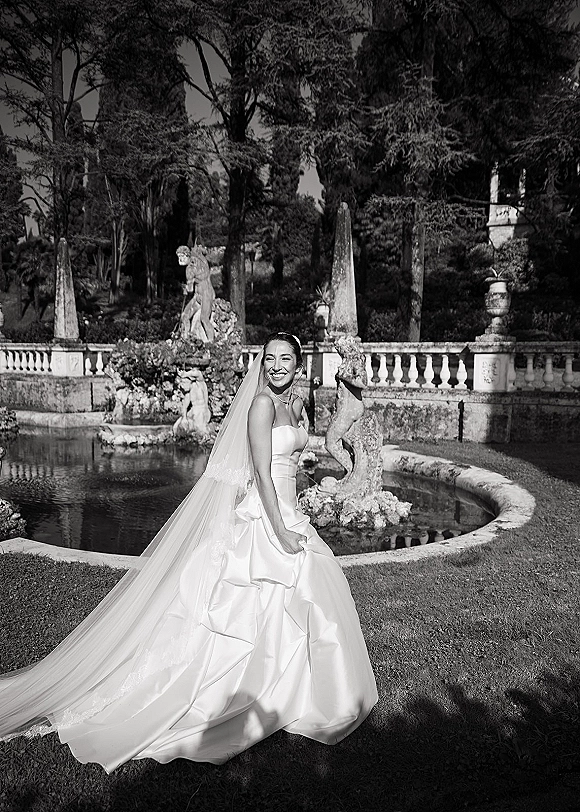 Bridal portrait of a smiling bride looking back in a strapless satin gown with a long veil, by a garden pond and stone fountain