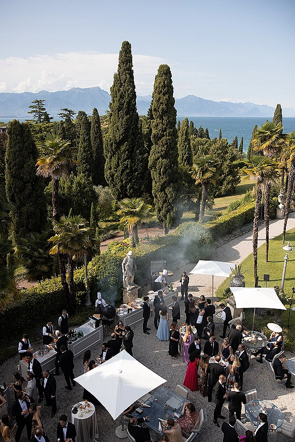 Wedding cocktail hour with outdoor wedding cocktail hour bar, high-top tables and umbrellas on a gravel terrace with lake and mountain views