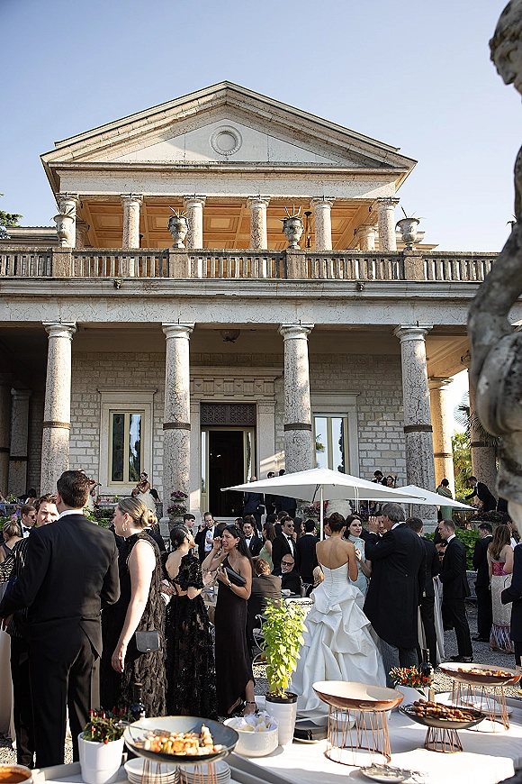 Wedding cocktail hour with guests mingling in formal attire around a buffet table under a white umbrella in a stone villa courtyard