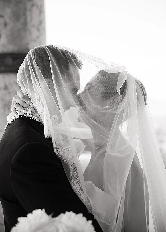 Wedding kiss portrait of bride and groom under a veil, lace edge framing their faces as bright window light softens the scene