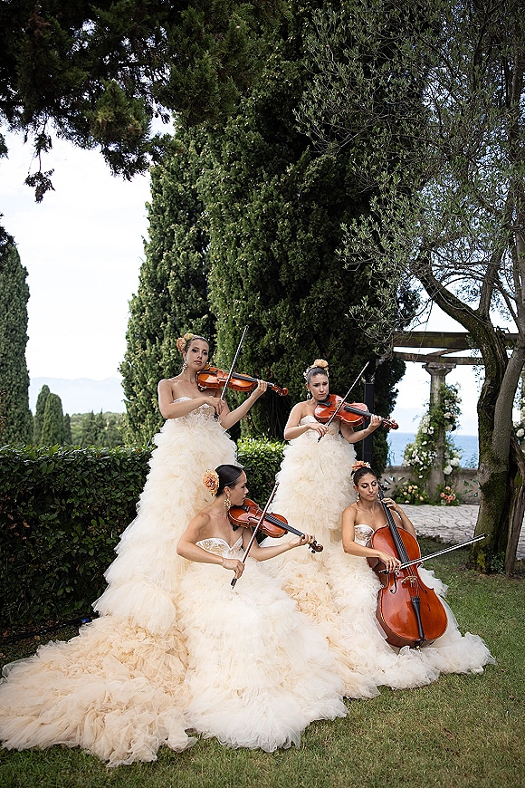 Wedding musicians in strapless ruffle gowns play violin and cello on a garden lawn, floral hair accents with pergola and hills behind