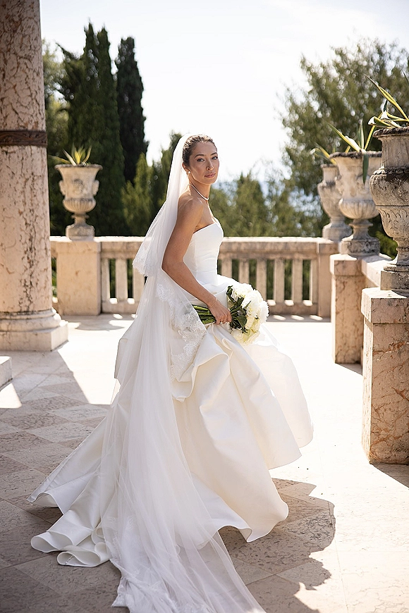 Bridal portrait of a bride in a strapless wedding dress with long veil, holding a white bouquet on a stone terrace with columns and balustrade
