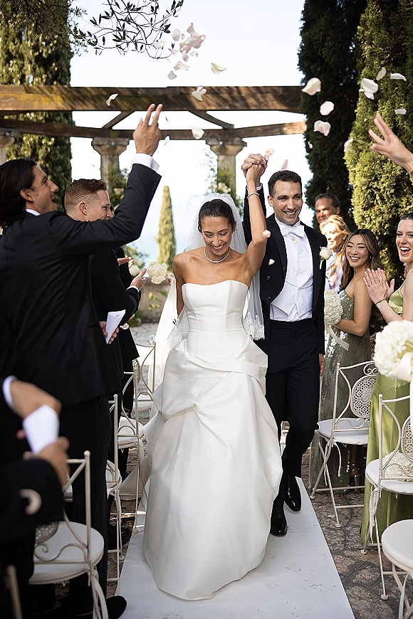 Ceremony recessional as bride and groom walk the aisle holding hands, petals tossed by guests in an outdoor garden pergola setting