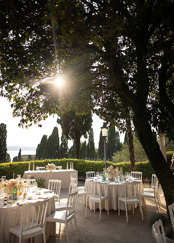 Outdoor reception setup with round tables, white linens, floral centerpieces, candle lanterns, and glassware on a sunlit stone patio by trees and water view