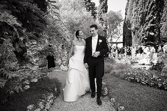 Wedding couple portrait of bride and groom walking hand in hand, bride holding bouquet with veil flowing along a garden path by a stone wall