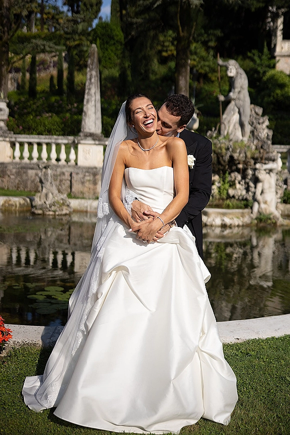 Couple portrait of bride and groom embrace as he kisses her cheek, cathedral veil and pearls in a garden by a pond with stone statues