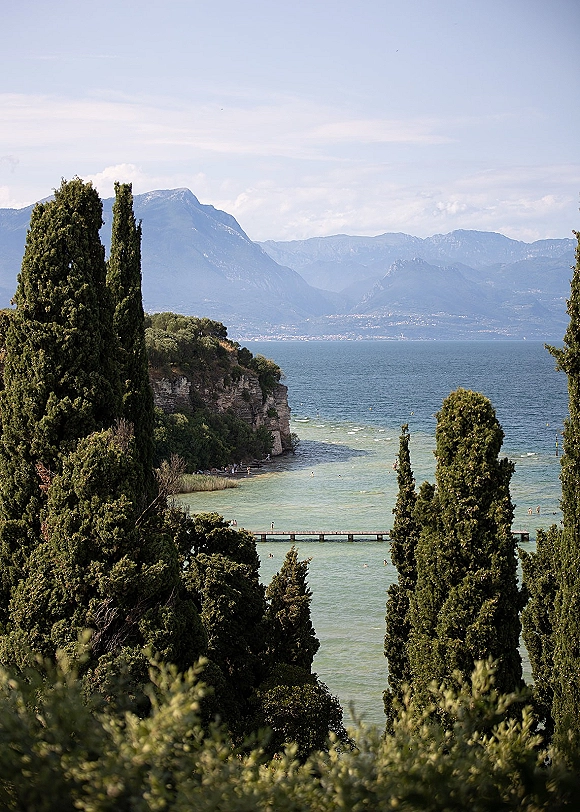 Lakeside landscape with lake view mountains, turquoise water and cypress trees lining a cliffside shoreline near a pier under open sky