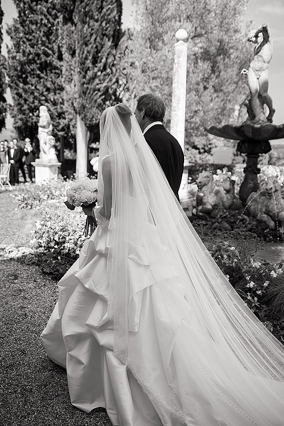 Bride and groom walking down the aisle, her long wedding veil and white rose bouquet trailing along a garden path by a stone fountain