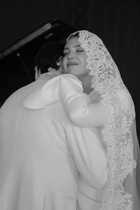 Wedding hug as bride and groom embrace closely, her eyes closed under a lace veil, his suit jacket framing a dark interior backdrop