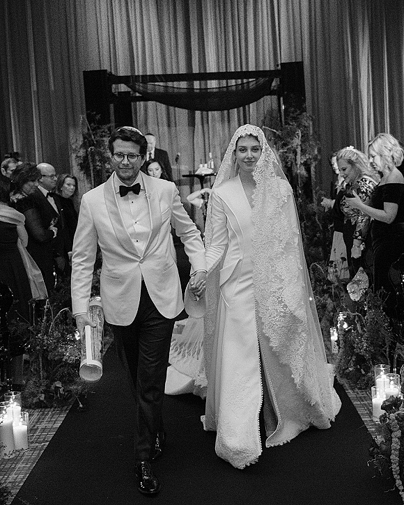 Wedding recessional as newlyweds walking aisle hand in hand, bride in lace veil and long sleeves, groom in white jacket, candlelit chuppah backdrop