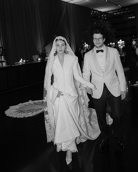 Newlywed couple holding hands during a bride and groom entrance, bride in lace veil and gown, groom in white tuxedo jacket at candlelit reception